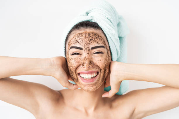 young smiling woman applying coffee scrub mask on face - happy girl having skin care spa day at home - healthy alternative natural exfoliation treatment and people lifestyle concept - pampering massaging indoors adult imagens e fotografias de stock