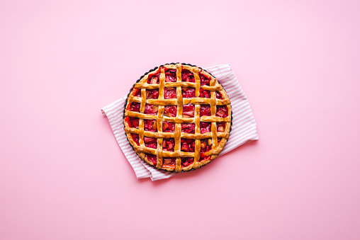 Homemade rhubarb and strawberries pie on a pink background, above view. Flatlay with a freshly baked lattice crust pie