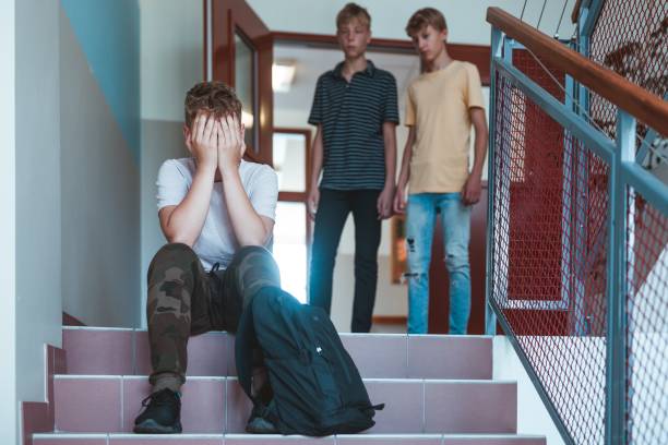 istock Sad kid with backpack sitting on stairs at school, two classmates behind him 1226815693