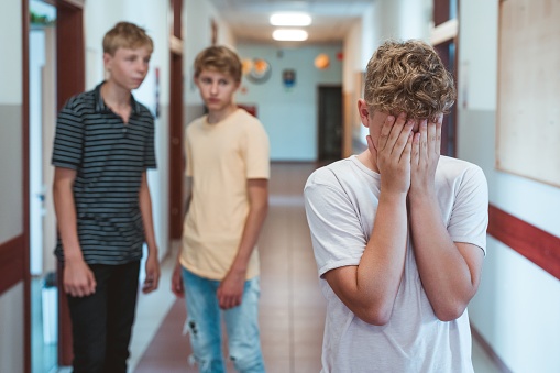 Sad bullied boy standing n the school corridor, covering his face. Two classmates behind him