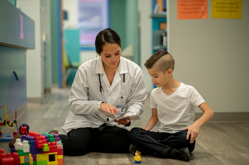 Young female speech therapist sits on floor with young male patient pointing to images in a book for him to enunciate.