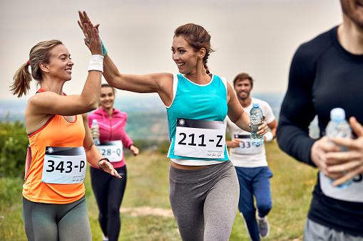 Happy athletic women running a marathon and giving high-five to each other during the race in nature.