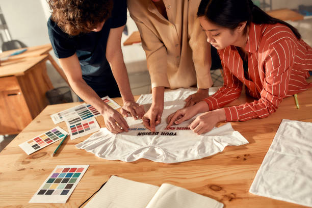 dove tecnologia e artigianalità si incontrano. persone creative che provano gli adesivi con testo, mentre discutono del design della t-shirt. giovani uomini e donne che lavorano presso t-shirt personalizzata, azienda di stampa di abbigliamento - textile print foto e immagini stock