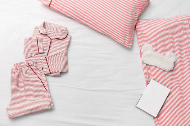 眠りのためのスリープウェア。ピンクの女性は、ストライプ、枕、ふわふわ睡眠アイマスクと寝る前に読書のための本と綿の布からパジャマ。トップ ビュー。フラットレイ。