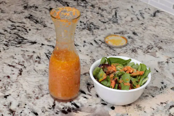 Freshly prepared carrot ginger salad dressing in a clear glass bottle next to a spring mix salad in a bowl. Healthy mixed greens salad in a bowl with carrot ginger salad dressing on a stone counter.
