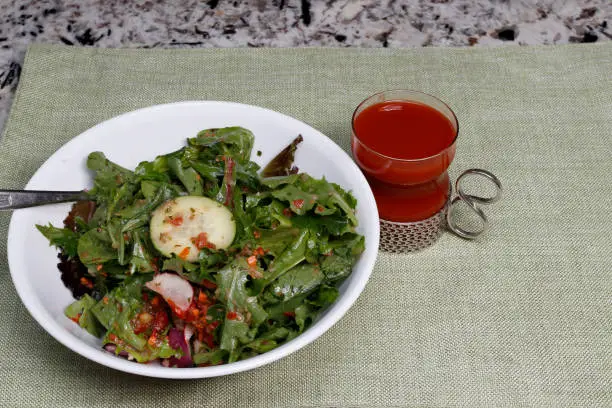 Healthy spring mix salad in a bowl with vegetables and carrot ginger salad dressing and tomato juice. Meal of mixed salad greens in a white bowl with a fork near a tomato beverage on placemat.
