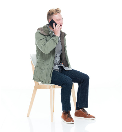 Profile view of aged 18-19 years old caucasian male sitting in front of white background wearing jeans who is talking and using mobile phone