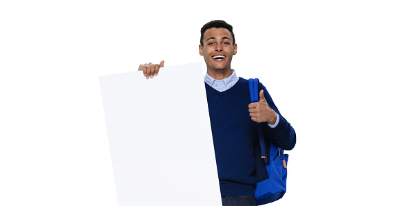 Full length of aged 20-29 years old with black hair generation z male standing in front of white background wearing warm clothing who is laughing and showing thumbs up who is showing with hand and holding banner sign with copy space