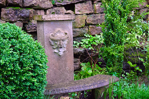 Remarkably vivid sculpted face of demon or green man on a rectangular column behind a stone bench, surrounded by lush greenery