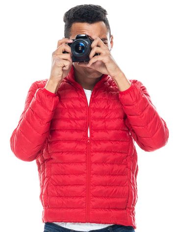 Front view of aged 20-29 years old with black hair african ethnicity male tourist standing in front of white background wearing jacket who is cheerful who is photographing and holding camera