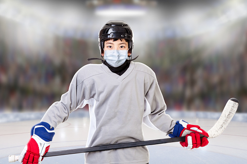 Junior ice hockey player with full equipment and sports uniform posing in fictitious arena with face mask. Concept of new normal in sports to prevent against Covid-19 coronavirus spread when sports leagues or competition resume.