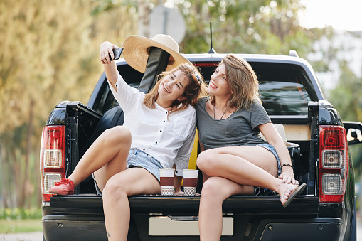 Cheerful traveling female friends sitting in car trunk and taking selfie