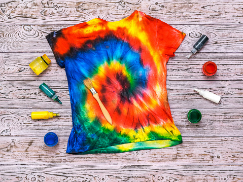 T-shirt painted in the style of tie dye with colors on a wooden background. White clothes painted by hand. Flat lay.