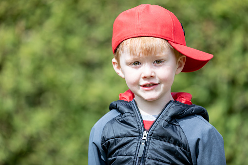 A little redhead kid is smiling and looking at camera. The little redhead boy is outdoors. It is a beautiful sunny day. He is wearing a red hat.