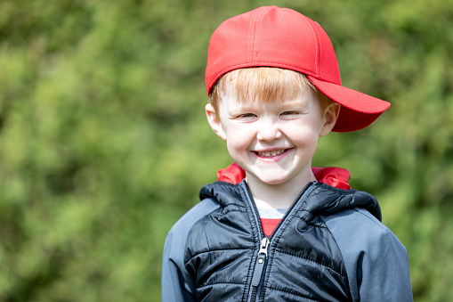 A little redhead kid is smiling and looking at camera. The little redhead boy is outdoors. It is a beautiful sunny day. He is wearing a red hat.