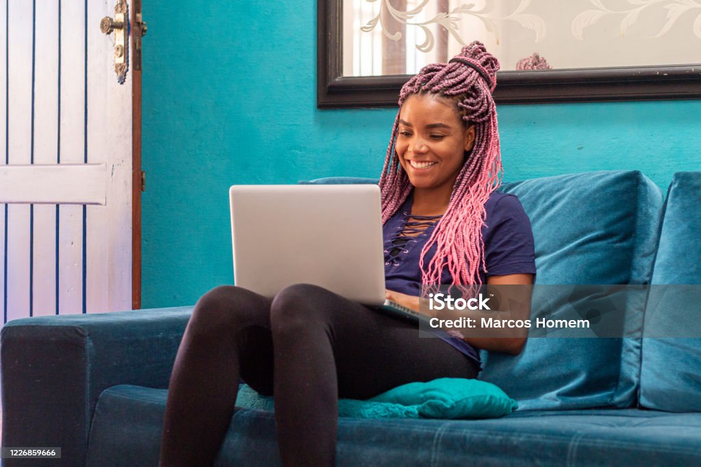 Young black woman with braid using notebook in home room Young black woman with pigtails using notebook sitting on blue sofa in home room Teenager Stock Photo