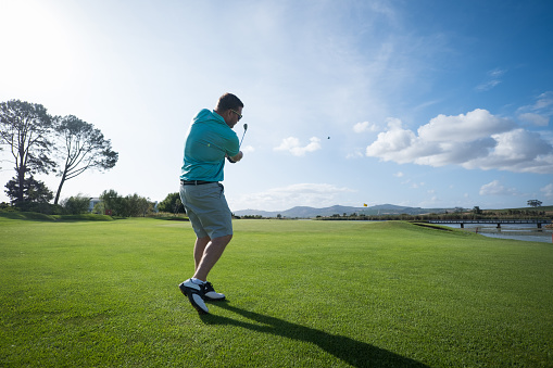Man playing golf on field. Three quarter length of player is wearing sports clothing. Golfer hitting his shot to a green.