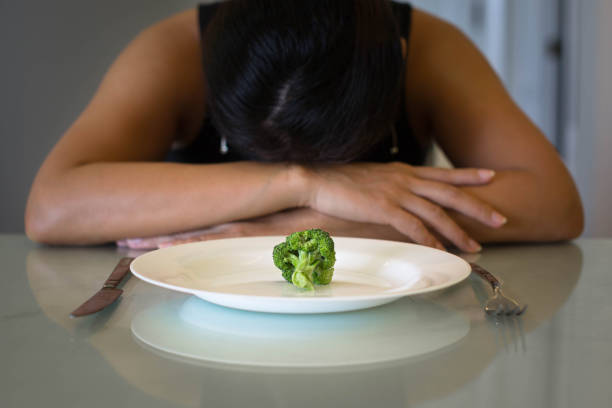 Depressed woman hungry from dieting, sitting in front of a empty plate. Weight loss diet. A woman sitting in front of a plate with a small portion of vegatables, looking miserable and hungry. bulimia stock pictures, royalty-free photos & images