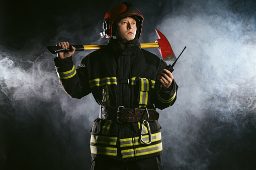 Portrait of a firefighter using holding a fire hose while his colleague is storing a chainsaw inside of a fire truck equipment compartment