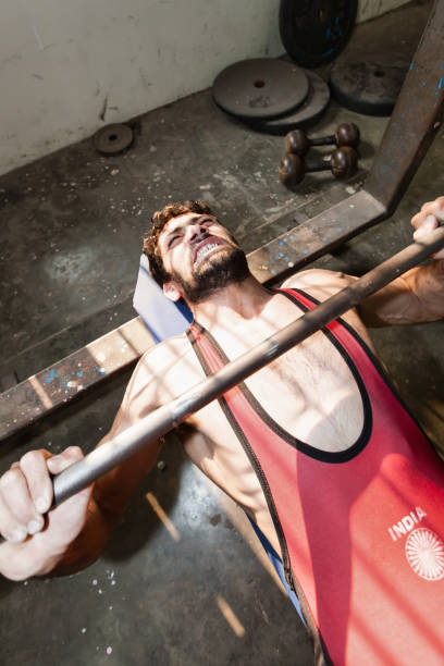 Indian Wrestler Lifting Weights New Delhi India Indian Wrestler training his muscles in old rotten training backyard room in downtown New Delhi. Pushing weights lying on his back on metal construction weight bench. The India Logo on his shirt is a generic part of the Indian National Flag. New Delhi, India, Asia. wrestlers uniform stock pictures, royalty-free photos & images