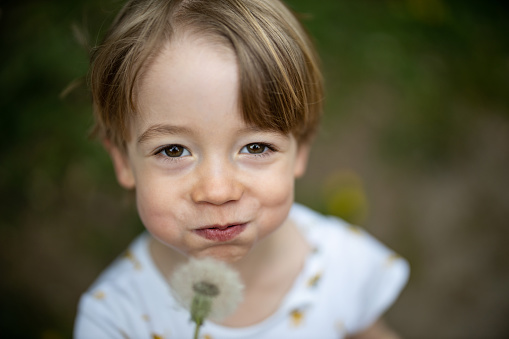 Young boy blowing a dandelion while smiling at the camera