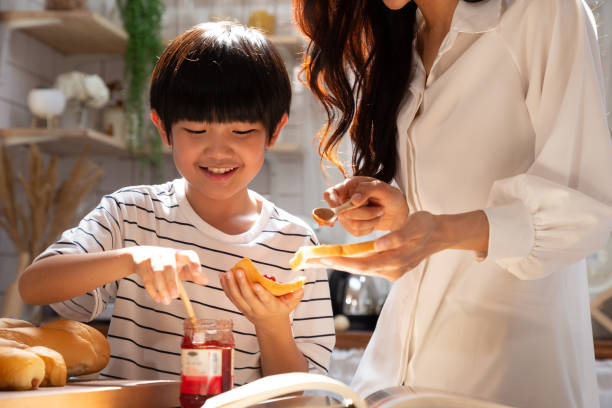 mãe e filho sorridentes cozinhando e fazendo comida de café da manhã com pão e geleia de morango juntos na cozinha de casa. conceito de atividade de vida feliz - healthy eating snack child domestic kitchen - fotografias e filmes do acervo