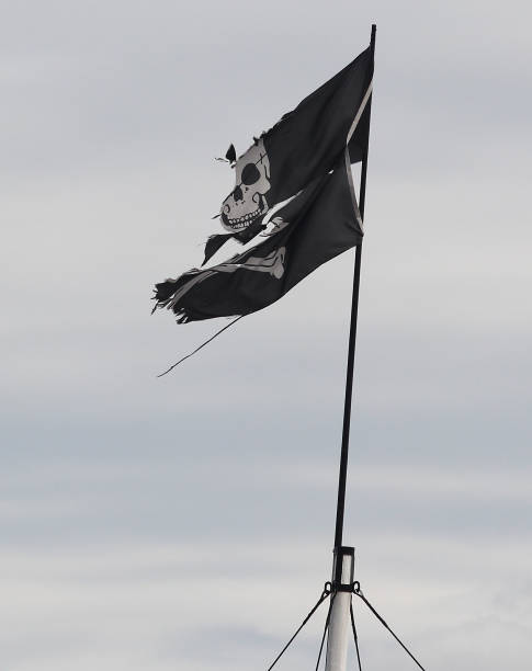 A torn & ripped Jolly Roger Pirate Flag A general view of a torn and ripped Jolly Roger Pirate flag flutters in the wind against an overcast grey sky. pirate flag stock pictures, royalty-free photos & images