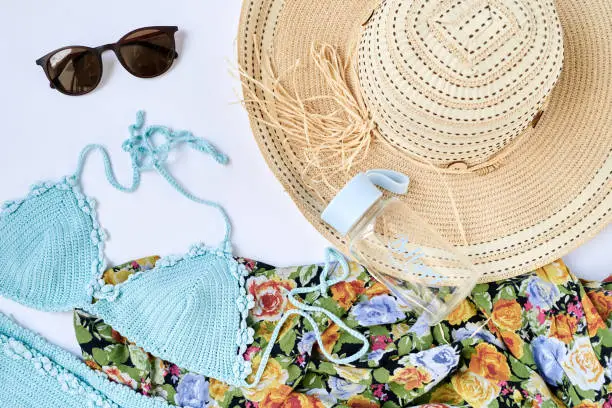 Flat lay picture of summer vacation beach accessories, isolated on white background. Mint light blue swimsuit, sunglasses, colorful flowery scarf, bottle of water with Be happy sign and straw hat.