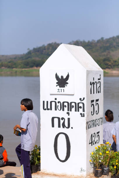 Thai border stone with km 0 at Mekong river at promenade in Chiang Kham area in Chiang Rai province. Behind stone are thai students in school uniform at waterfront. In background are river Mekong and Laos.