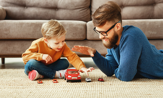 Side view of positive young bearded man and little son sitting on floor near sofa and playing the toys cars  while enjoying time together at home