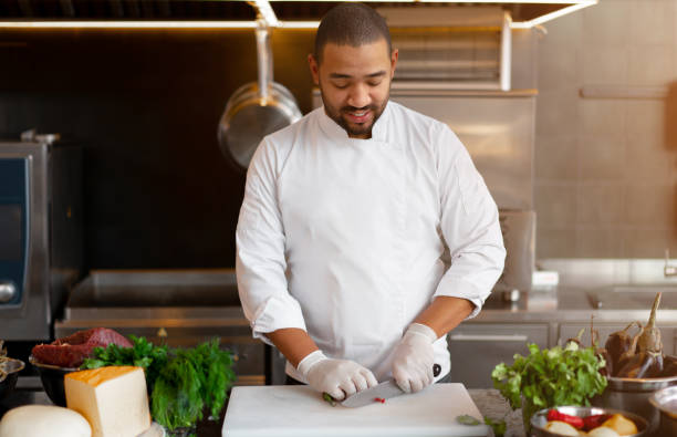 istock Handsome young African chef standing in professional kitchen in restaurant preparing a meal of meat and cheese vegetables. 1226943276