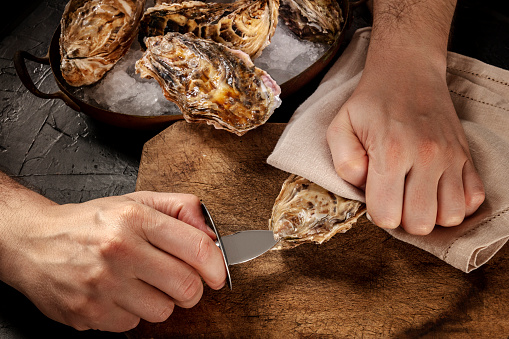 Shucking an oyster, man's hands with a special knife, opening oysters on a wooden board