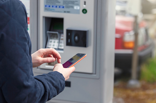 Closeup of a person paying a parking ticket with the mobile phone, next to a parking ticket machine at a parking lot.