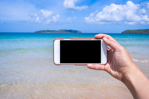 Tourist using phone at the beach with the sea, hand holding white mobile smart phone smartphone, travel working concept, blurry background, close up.