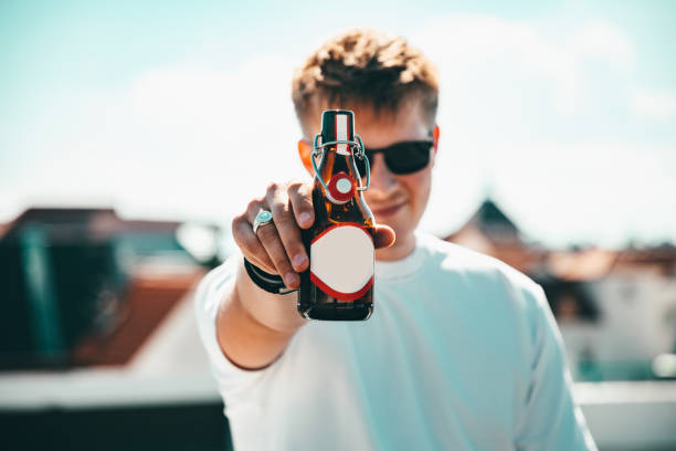 Beer o'Clock Young Man Inviting to a bottle of beer Young man standing on rooftop in summer wearing sunglasses passing over a bottle of beer during a rooftop party. Smiling happy and friendly. Youth Culture Summer Lifestyle Concept Shot. beer bottle photos stock pictures, royalty-free photos & images