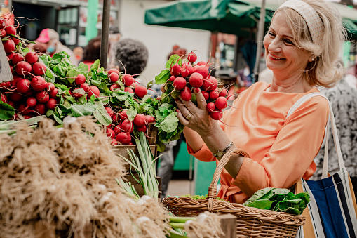 A happy smiling senior woman is holding a basket full of groceries on a farmers market