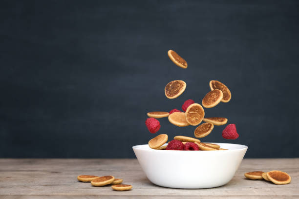 fliegenkleine pfannkuchen müsli - trendige quarantäne-lebensmittel. mini pfannkuchen und frische himbeeren in bewegung fallen in weißer schüssel auf schwarzetafel hintergrund. levitation, schwerkraft. hausgemachtes frühstück. - falling fruit berry fruit raspberry stock-fotos und bilder