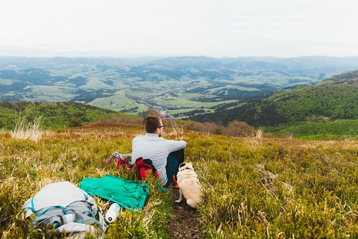 Young man hiker and his small fluffy cute dog - pug breed relaxing on the bright colorful meadow with panoramic view of the scenic Carpathian mountains, Ukraine