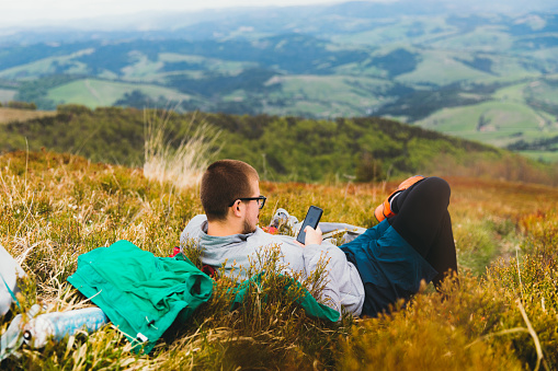 Young man in eyeglasses resting on the colorful meadow with panoramic view of the beautiful mountain peaks and texting message to his friends in Carpathian Mountains, Ukraine