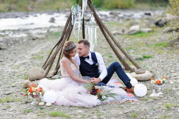 An attractive newlywed couple, a happy and joyful moment. Man and woman hug and kiss in holiday clothes. Wedding Cermonia with Boho-style décor by the river in the fresh air.
