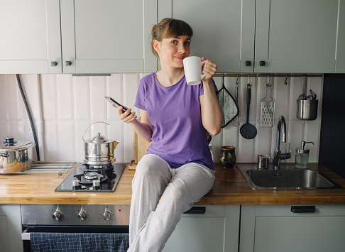 Young woman in the kitchen, smiling while drinking from a mug and using mobile phone