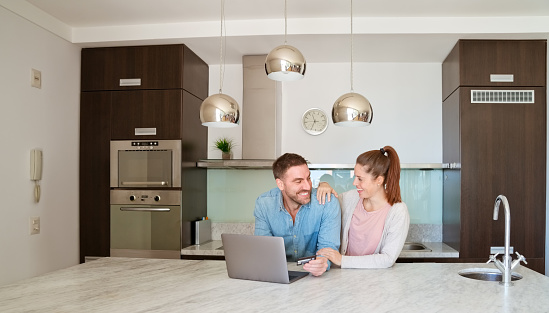 Women and man sitting at the table in the kitchen at home and doing online shopping. Adult man using laptop and holding credit card. Smiling woman embracing him.