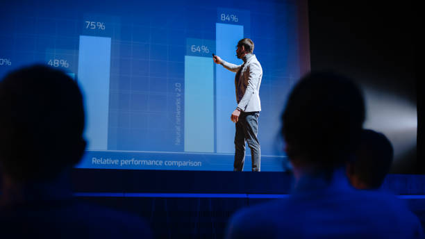 on stage visionary male speaker presents new technology, uses remote control for presentation, showing infographics, statistics animation on screen. live event / start-up business conference - financial figures audio imagens e fotografias de stock