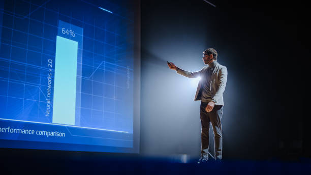 istock On-Stage: Handsome Speaker Does Presentation, New Product Release, Shows Infographics, Statistic Animation on Big Screen. Auditorium Hall Live Event, Start-up Conference, Device Presentation. 1226991401