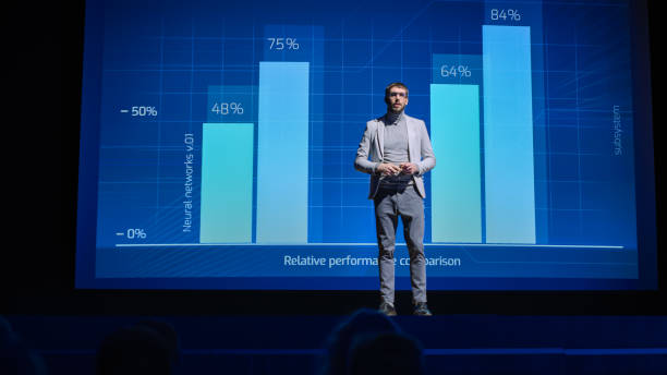 istock On-Stage: Speaker Does Presentation of the New Electronic Product, Shows Infographics, Statistic Animation on Screen. Auditorium Hall Live Event, Start-up Conference, Device Presentation and Release 1226991416