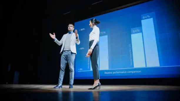 Photo of On Stage, Successful Female CEO and Male COO Speakers Present Company's New Product, Show Infographics, Statistics on Big Screen, Talk About Growth. Live Event, Tech Startup, Business Conference