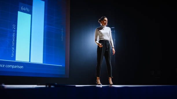 istock On-Stage Successful Female Speaker Presents Technological Product, Uses Remote Control for Presentation, Showing Infographics, Statistics Animation on Screen. Live Event / Device Release. 1226991423