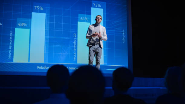 istock On-Stage: Speaker Does Presentation of the New Electronic Product, Shows Infographics, Statistic Animation on Screen. Auditorium Hall Live Event, Start-up Conference, Device Presentation and Release 1226991437