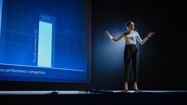 on-stage successful female speaker presents technological product, uses remote control for presentation, showing infographics, statistics animation on screen. live event / device release. - mobiles internet imagens e fotografias de stock