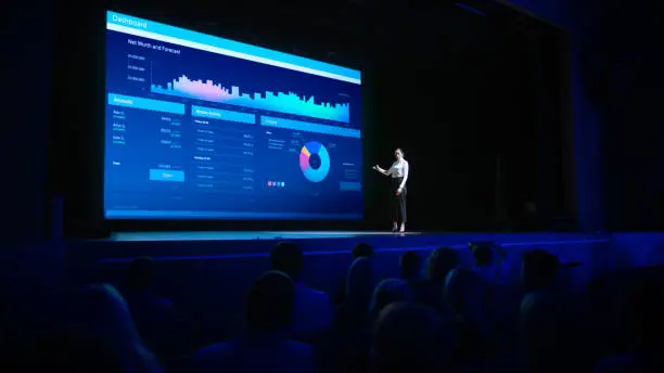 Business Forum Economics Conference Stage: Visionary Female Chief Analyst Delivers Speech and Shows Infographics, Statistics on Movie Theater Screen. Presentation with Speaker in Full Auditorium Hall